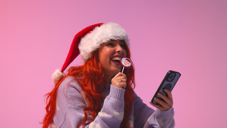 Studio-Shot-Of-Young-Gen-Z-Woman-Wearing-Christmas-Santa-Hat-Eating-Candy-Lollipop-Looking-At-Mobile-Phone-1